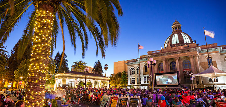 A palm tree trunk wrapped with lights in the foreground, and a crowd of people sitting in folding chairs facing a screen and a large building (Redwood City Courthouse) in the background.